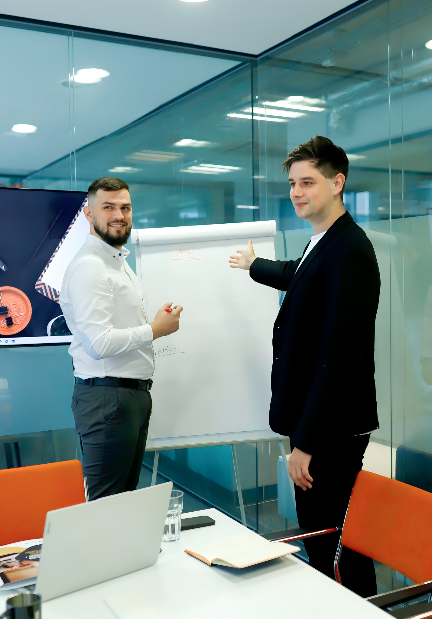 Dwóch mężczyzn w biurze prezentuje strategię na flipcharcie. Jeden wskazuje na napis, drugi trzyma marker. Widoczny laptop i szklanka wody na stole.