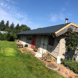 Sebastian Szymczak SzymBud - Budynek z szarej cegły z nowym, ciemnym dachem i widoczną membraną dachową. Czerwone drzwi, zielony trawnik i błękitne niebo w tle. Widoczne materiały budowlane.