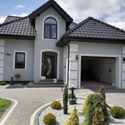 Sebastian Szymczak SzymBud - Szary dom jednorodzinny z czarnym dachem i elementami dekoracyjnymi, widok z przodu, z otwartym garażem i zadbanym ogrodem. Widoczne detale elewacji i oświetlenie zewnętrzne.