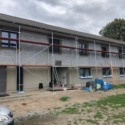 Sebastian Szymczak SzymBud - Budynek w trakcie ocieplania, widoczne rusztowania i materiały izolacyjne. Prace elewacyjne w toku, z detalami wykończenia okien i drzwi.