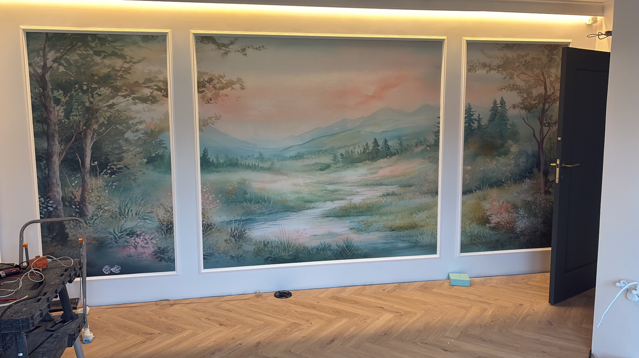 Trzyczęściowy mural z motywem pejzażu leśnego, otoczony białymi ramami, na ścianie w pokoju z parkietem ułożonym w jodełkę. Widoczny fragment drabiny malarskiej.