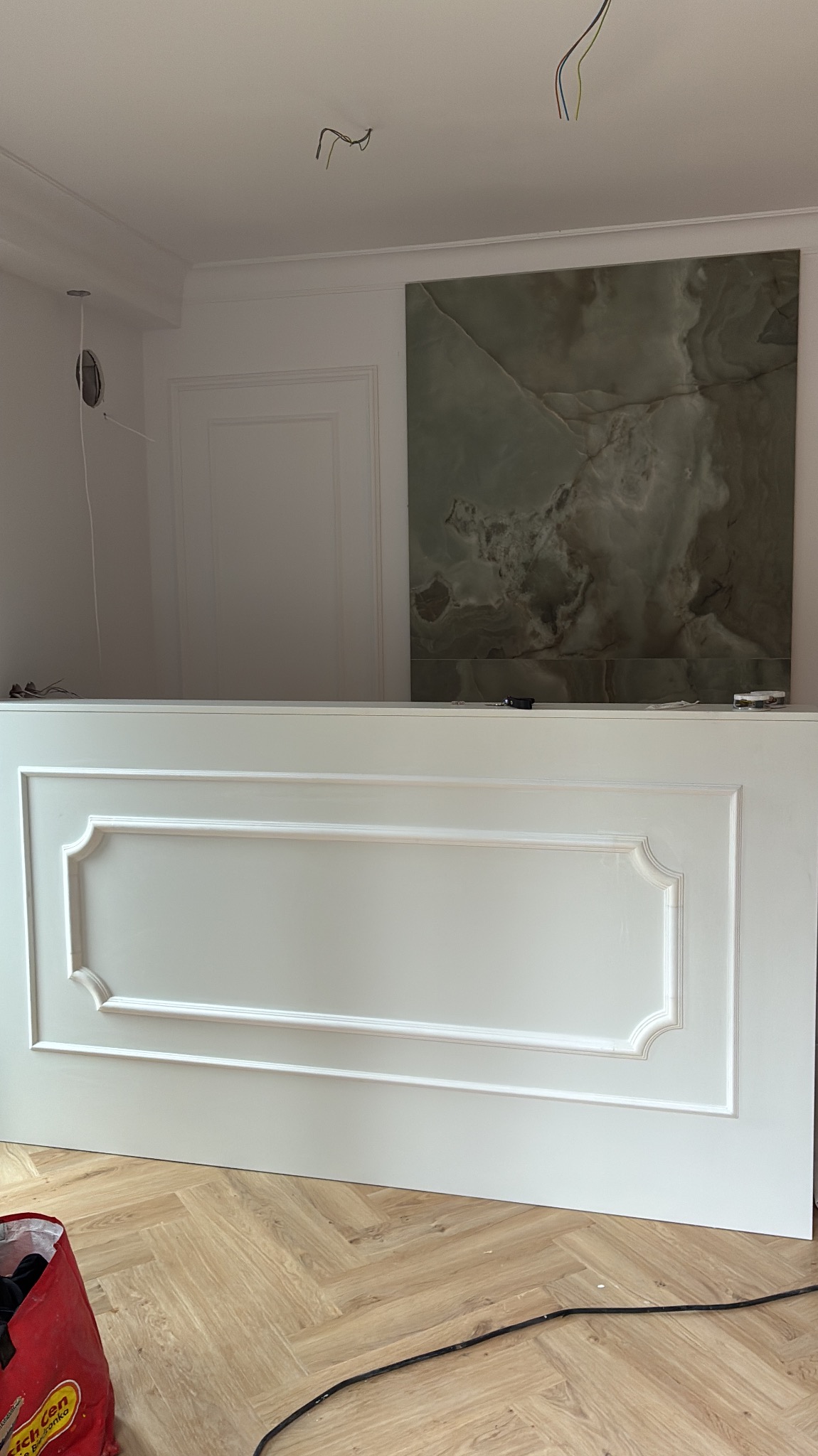 Biała, dekoracyjna boazeria z ozdobnym frezem na tle ściany z zielonym marmurem. Widoczne przewody elektryczne i parkiet w jodełkę. Detal wykończenia wnętrza.