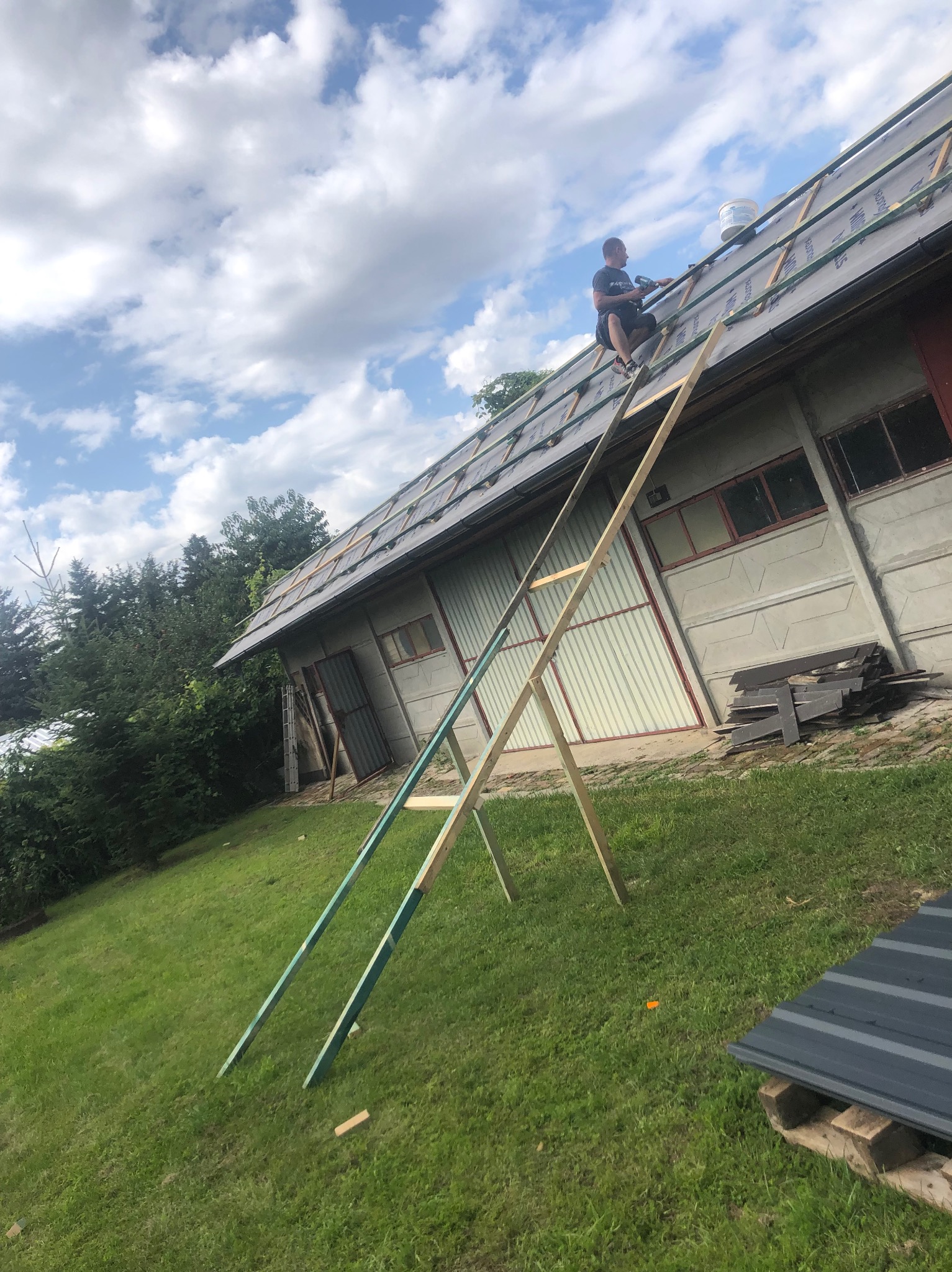 Pracownik na drabinie montuje pokrycie dachu budynku gospodarczego. Widoczne narzędzia i materiały budowlane. Ujęcie z dołu, na tle błękitnego nieba z chmurami.