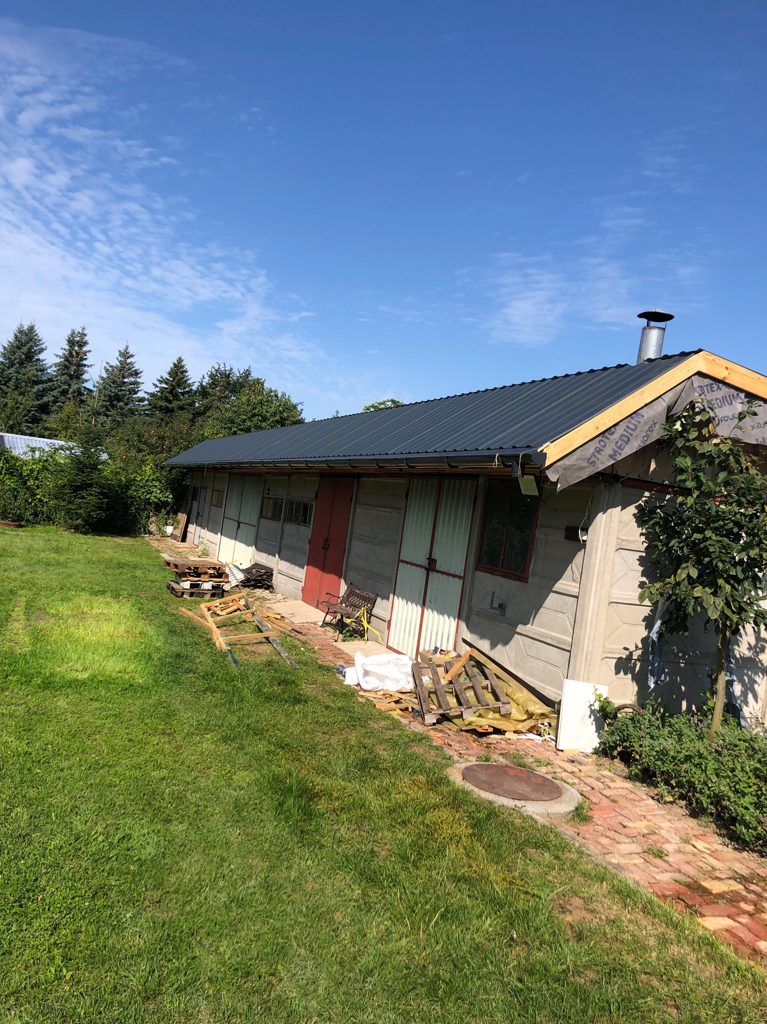 Budynek z szarej cegły z nowym, ciemnym dachem i widoczną membraną dachową. Czerwone drzwi, zielony trawnik i błękitne niebo w tle. Widoczne materiały budowlane.