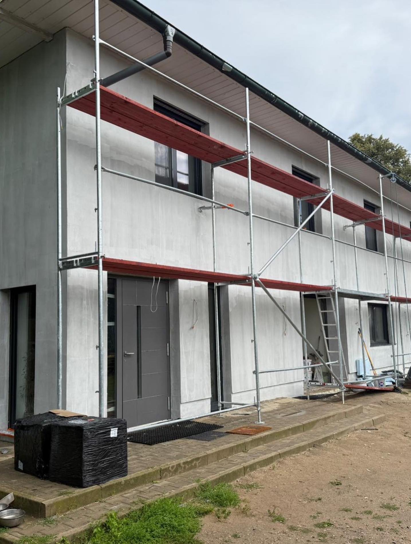 Dwukondygnacyjny dom w trakcie szpachlowania elewacji, z widocznymi rusztowaniami i materiałami budowlanymi. Szara elewacja, czarne okna i drzwi.