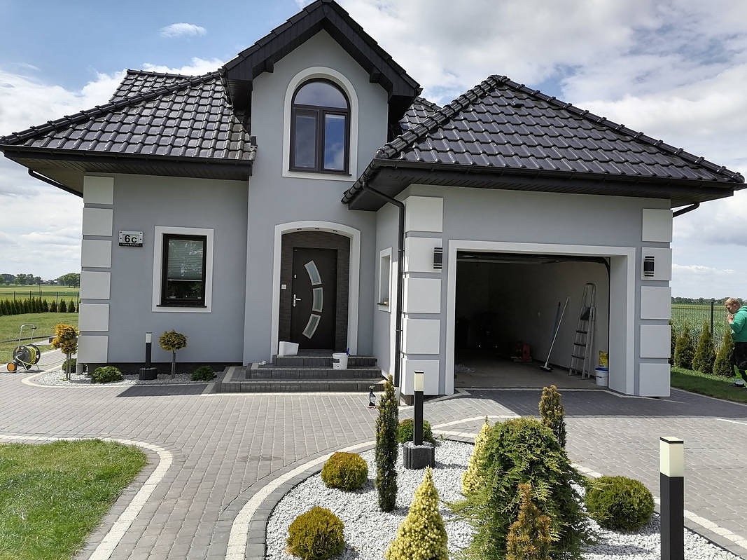 Szary dom jednorodzinny z czarnym dachem i elementami dekoracyjnymi, widok z przodu, z otwartym garażem i zadbanym ogrodem. Widoczne detale elewacji i oświetlenie zewnętrzne.