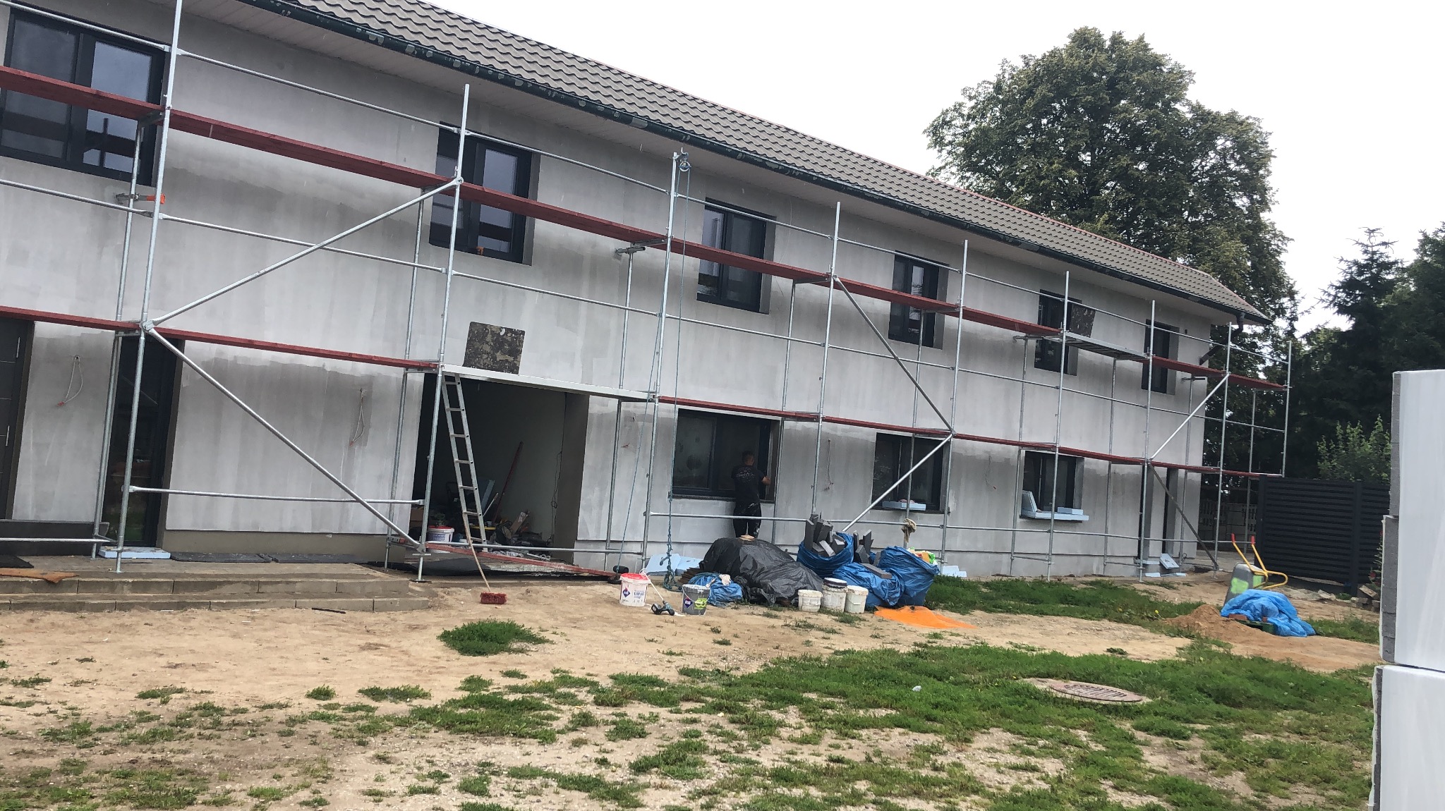 Dwukondygnacyjny dom w trakcie szpachlowania elewacji, z widocznymi rusztowaniami i materiałami budowlanymi na posesji. Widoczna osoba przy oknie.