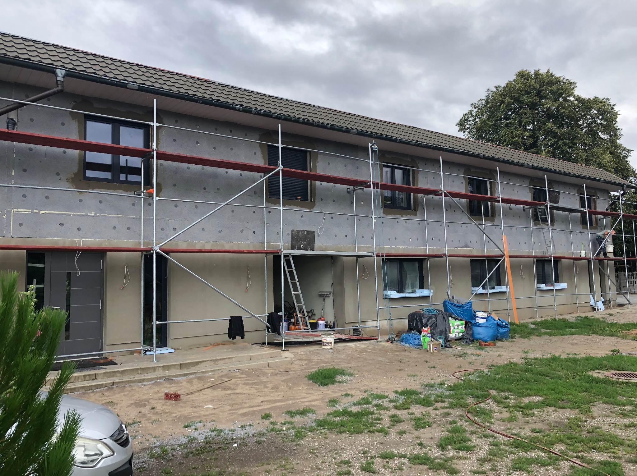 Budynek w trakcie ocieplania, widoczne rusztowania i materiały izolacyjne. Prace elewacyjne w toku, z detalami wykończenia okien i drzwi.