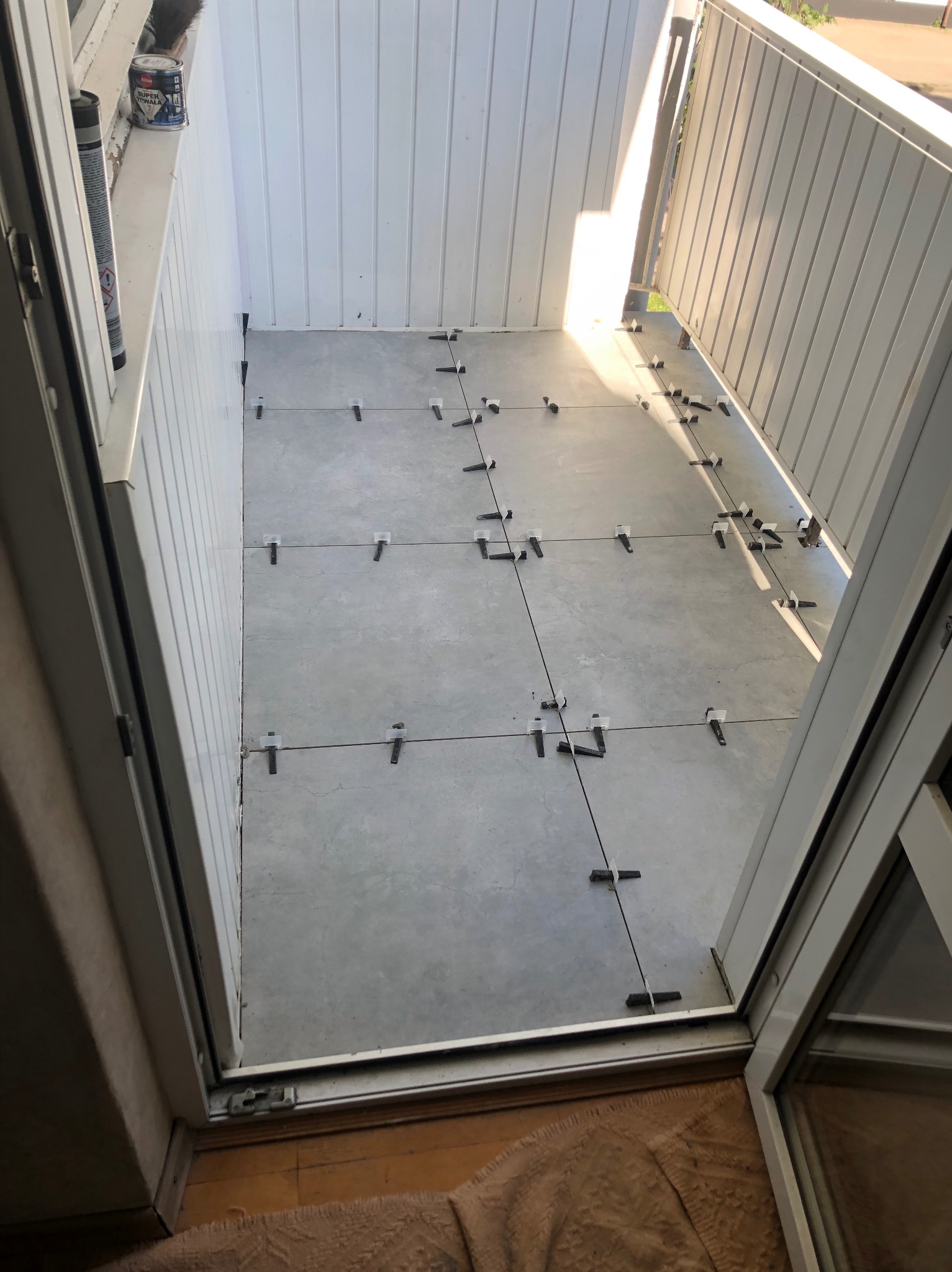 Jasne płytki ułożone na balkonie z użyciem krzyżyków dystansowych, widok z wnętrza mieszkania przez otwarte drzwi balkonowe. Świeże wykończenie.