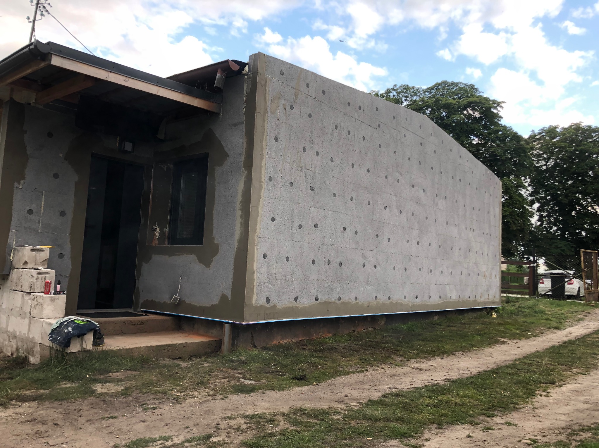 Dom w trakcie ocieplania: szara elewacja z widocznymi punktami mocowania izolacji, częściowo otynkowana, z widocznymi schodami wejściowymi i elementami otoczenia.