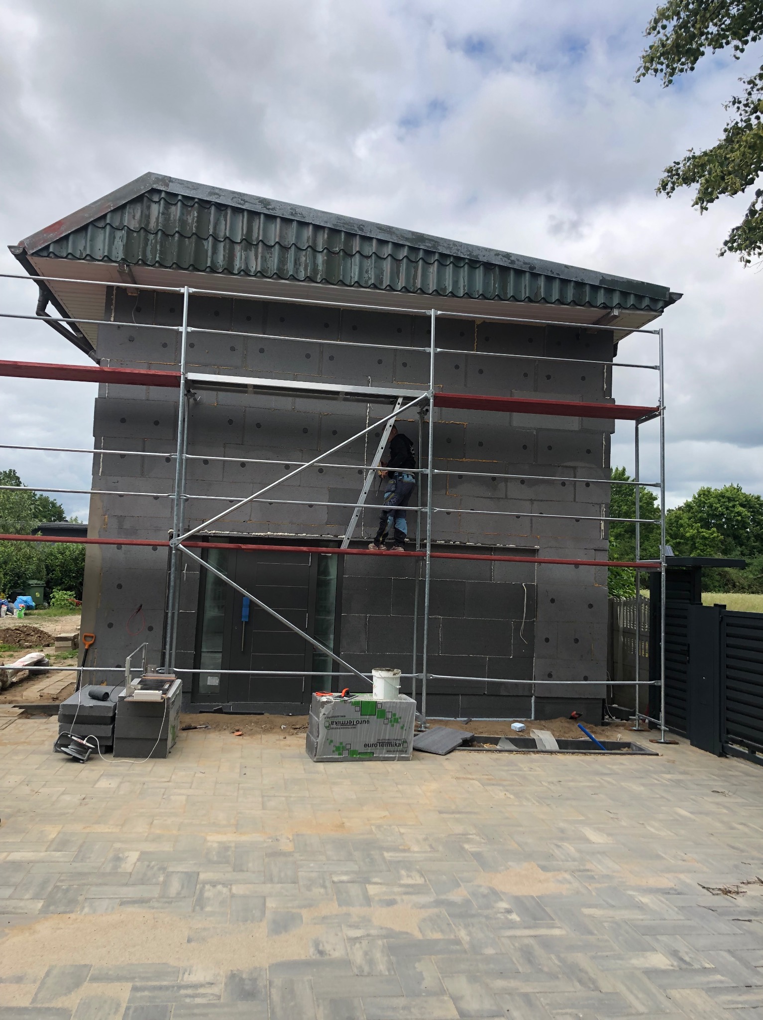 Ocieplanie elewacji budynku z szarego styropianu, widoczne rusztowanie i materiały budowlane na kostce brukowej. Prace w toku, dzień pochmurny.