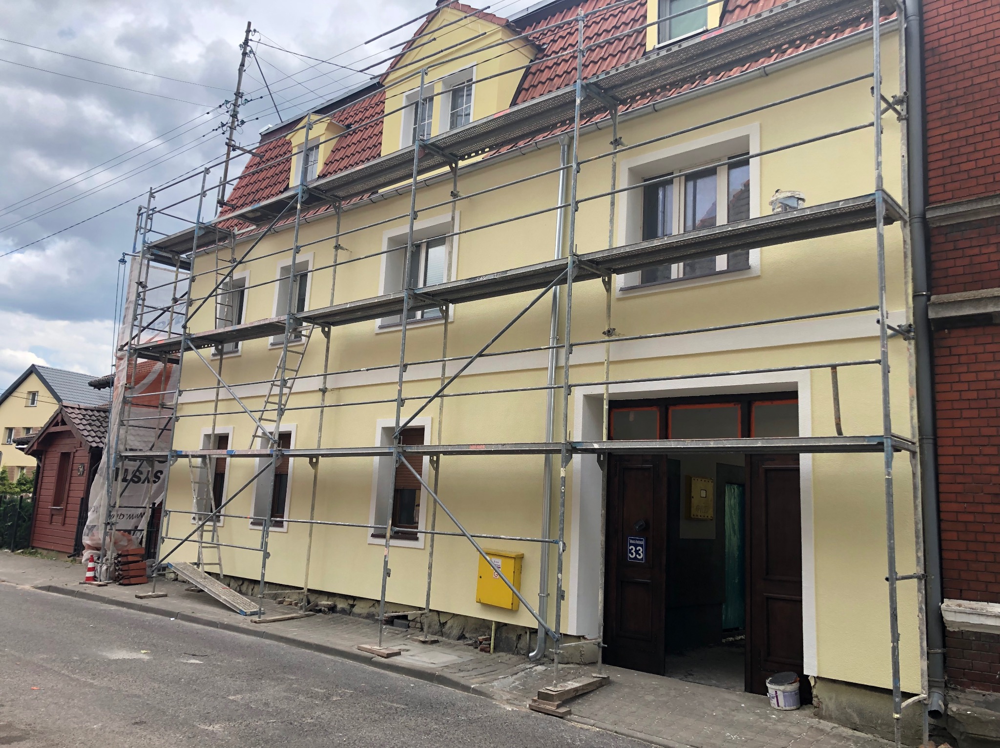 Żółta elewacja domu w rusztowaniach, widoczne detale architektoniczne i numer 33 nad wejściem. Świeżo pomalowana fasada, częściowo zasłonięta rusztowaniem.