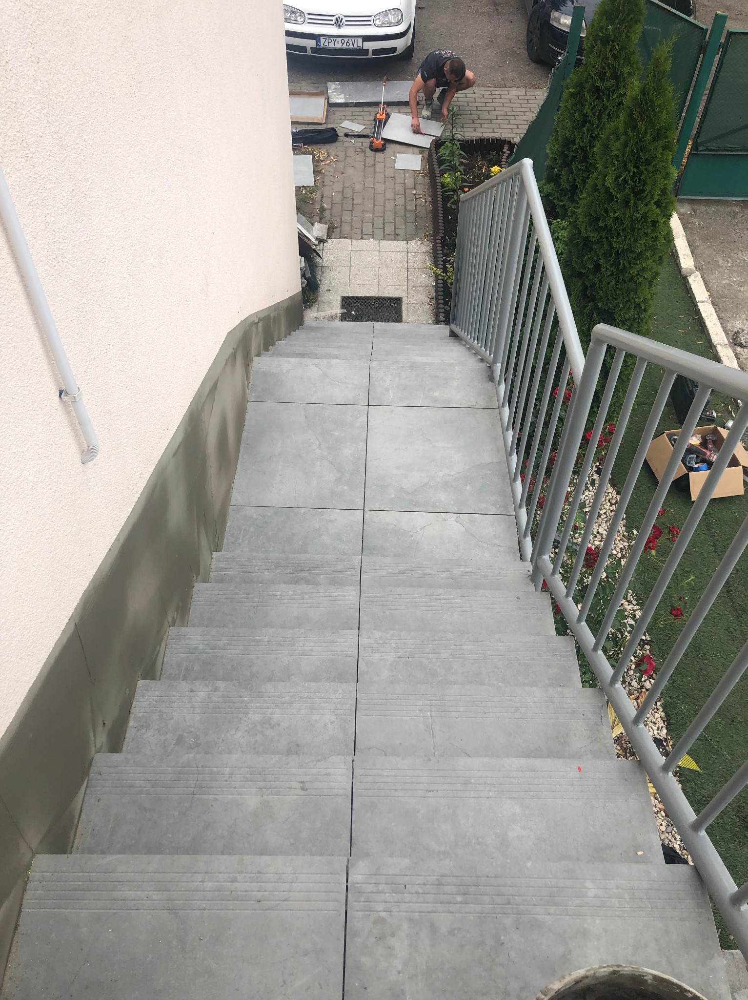 Wykończone schody zewnętrzne z szarych płyt, widok z góry. Metalowa barierka po prawej stronie. W tle osoba układająca płytki na chodniku.