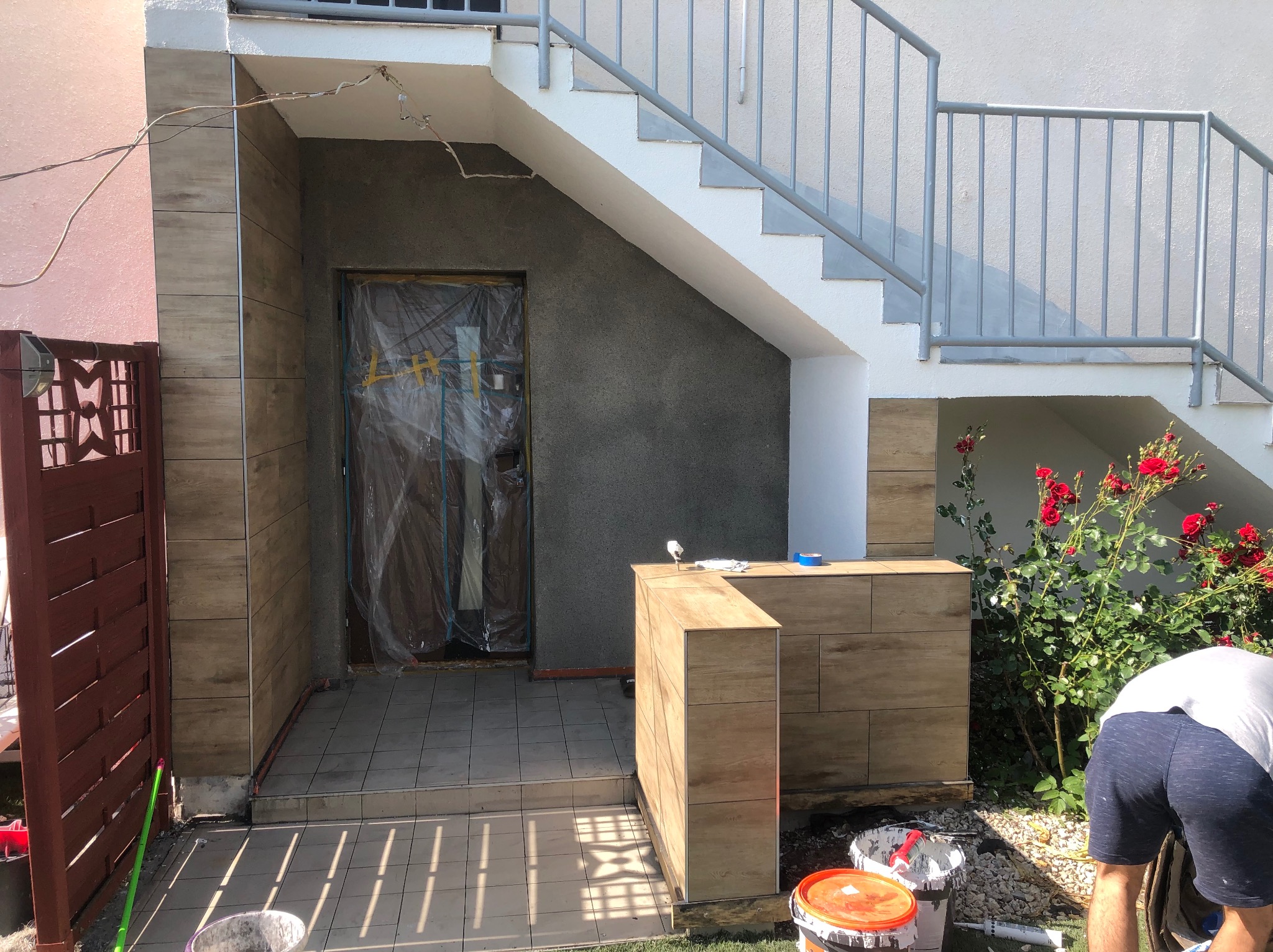 Wykończenie elewacji wejściowej z płytkami imitującymi drewno. Częściowo zakryte drzwi folią ochronną, widoczne schody i narzędzia pracy.