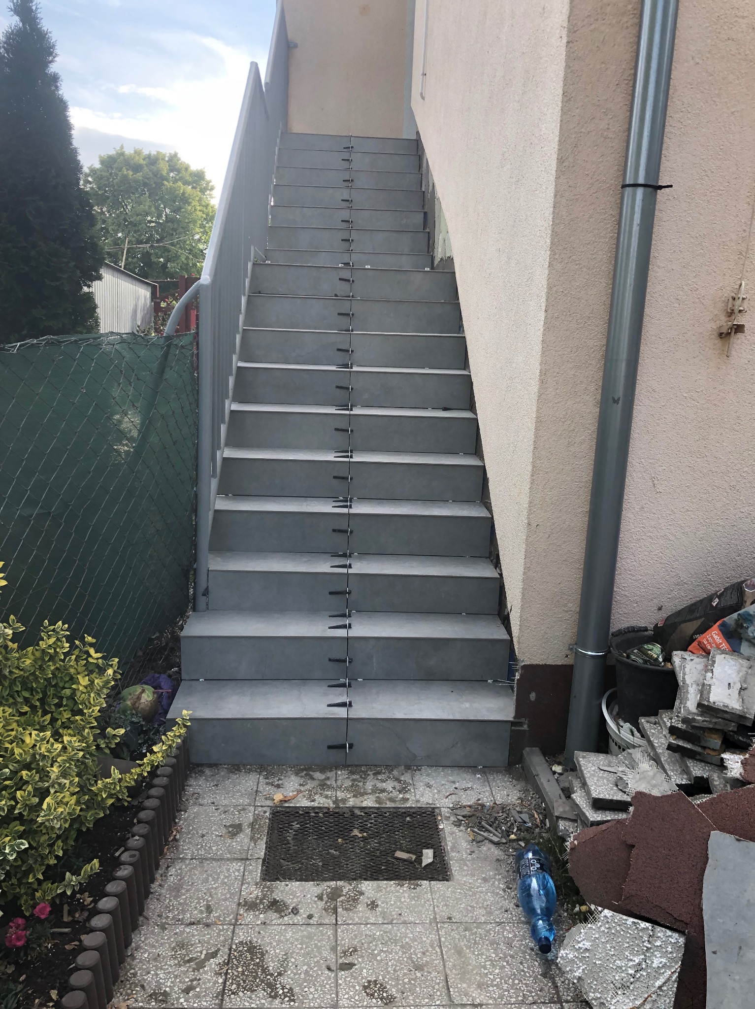 Zewnętrzne schody z szarych płyt, zabezpieczone czarnymi elementami montażowymi, prowadzące do budynku z beżową elewacją i szarą rurą spustową. Widoczny fragment ogrodzenia.