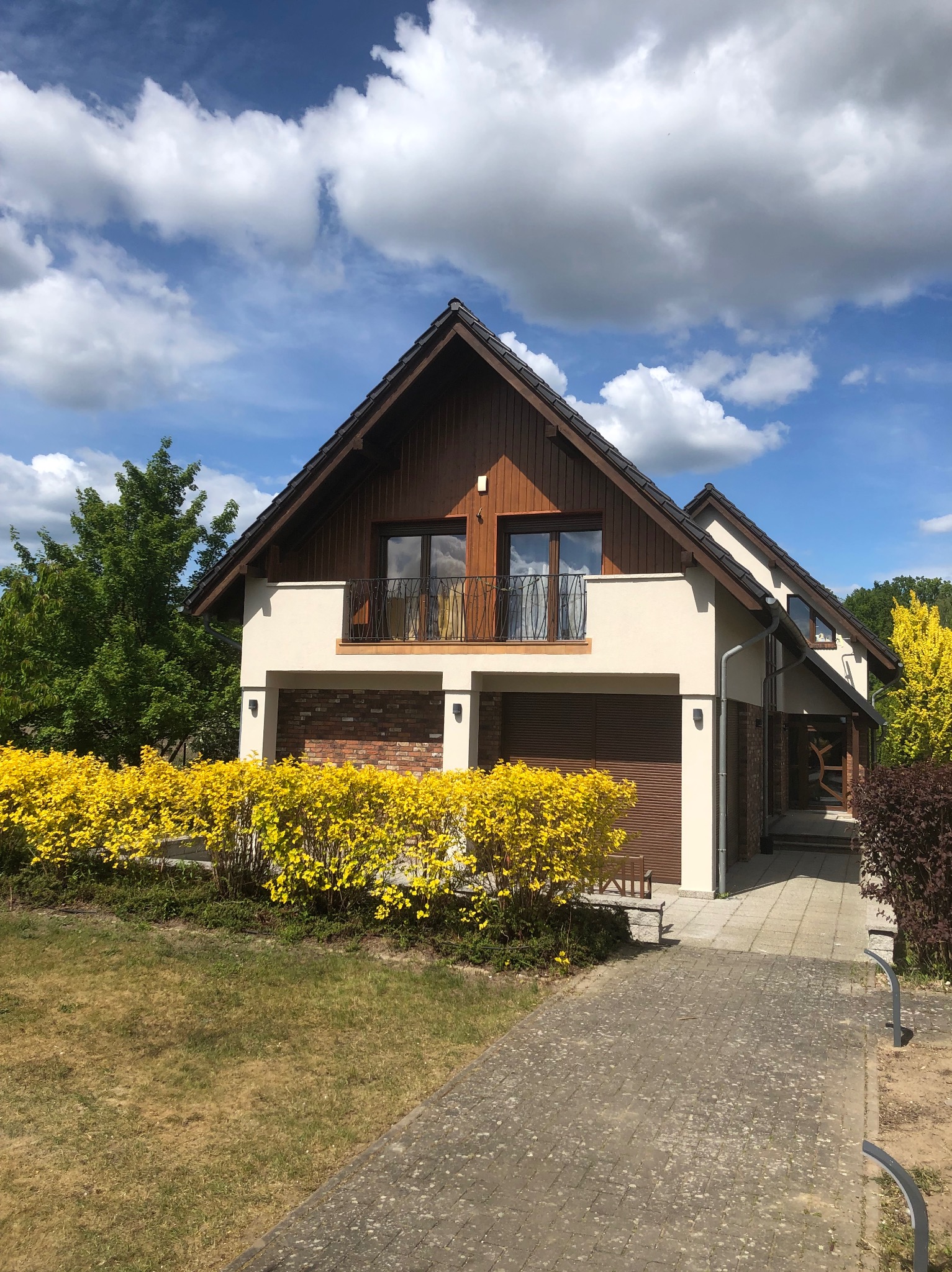 Dwukondygnacyjny dom z brązowym dachem, beżową elewacją i balkonem z kutą balustradą. Żółte krzewy przed budynkiem. Ujęcie w słoneczny dzień.