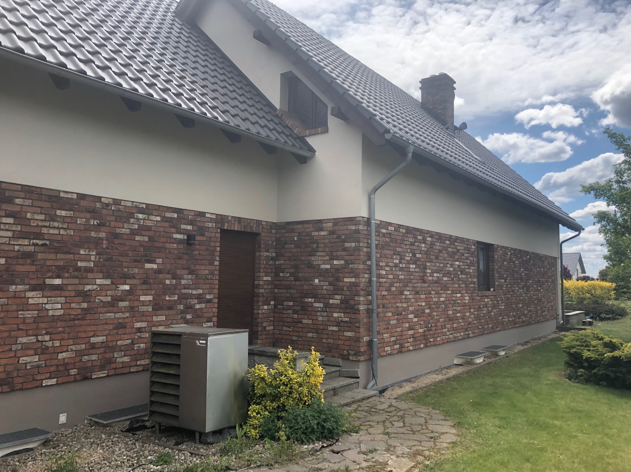 Elewacja domu jednorodzinnego z cegły klinkierowej, jasna góra, szara dachówka, widoczne rynny i fragment ogrodu. Ujęcie z poziomu trawnika, w tle błękitne niebo.