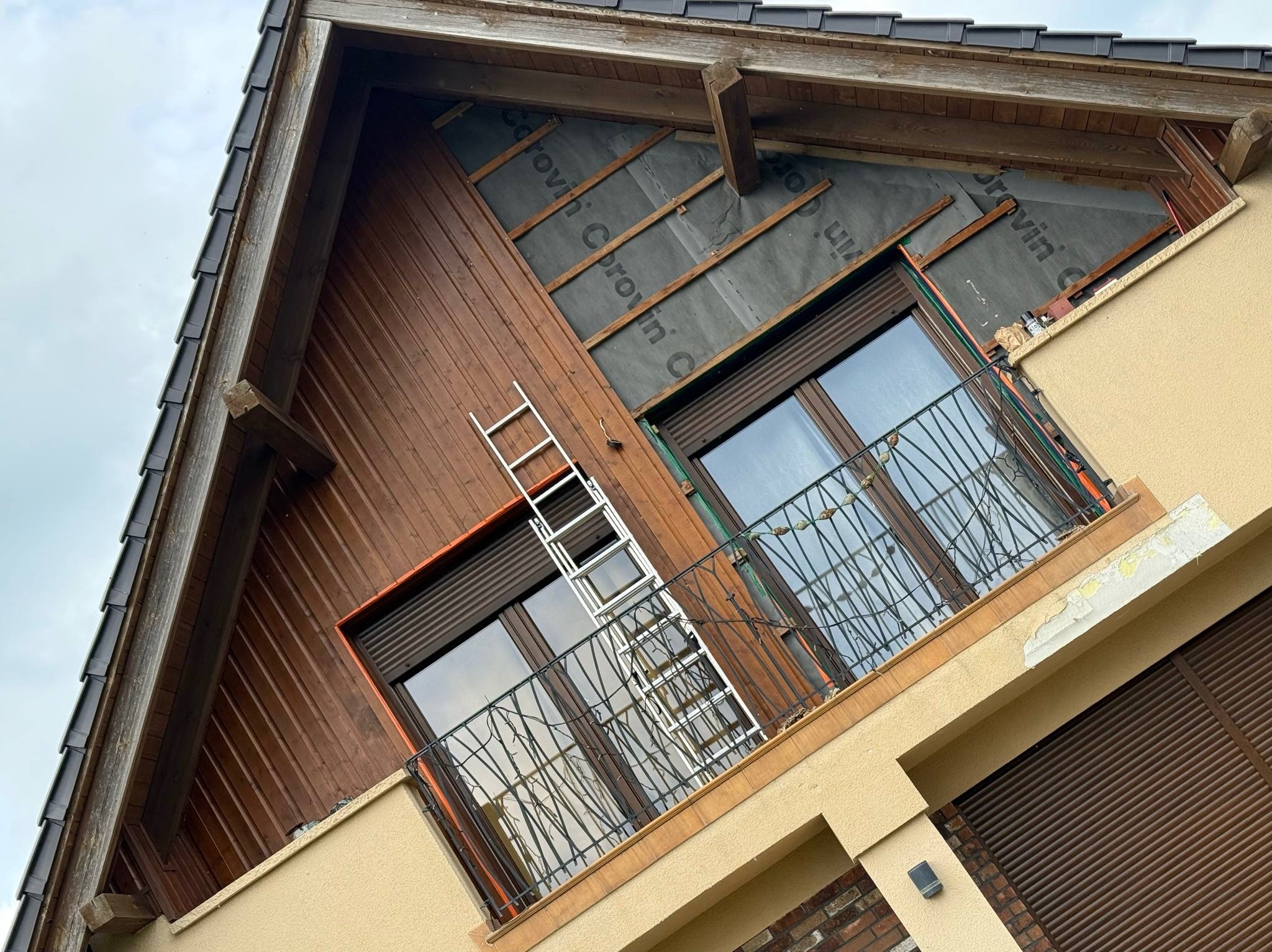 Drewniana elewacja domu w trakcie montażu, widoczna membrana i ruszt. Metalowa drabina oparta o balkon z ozdobną balustradą. Ujęcie z dołu, ukośne.