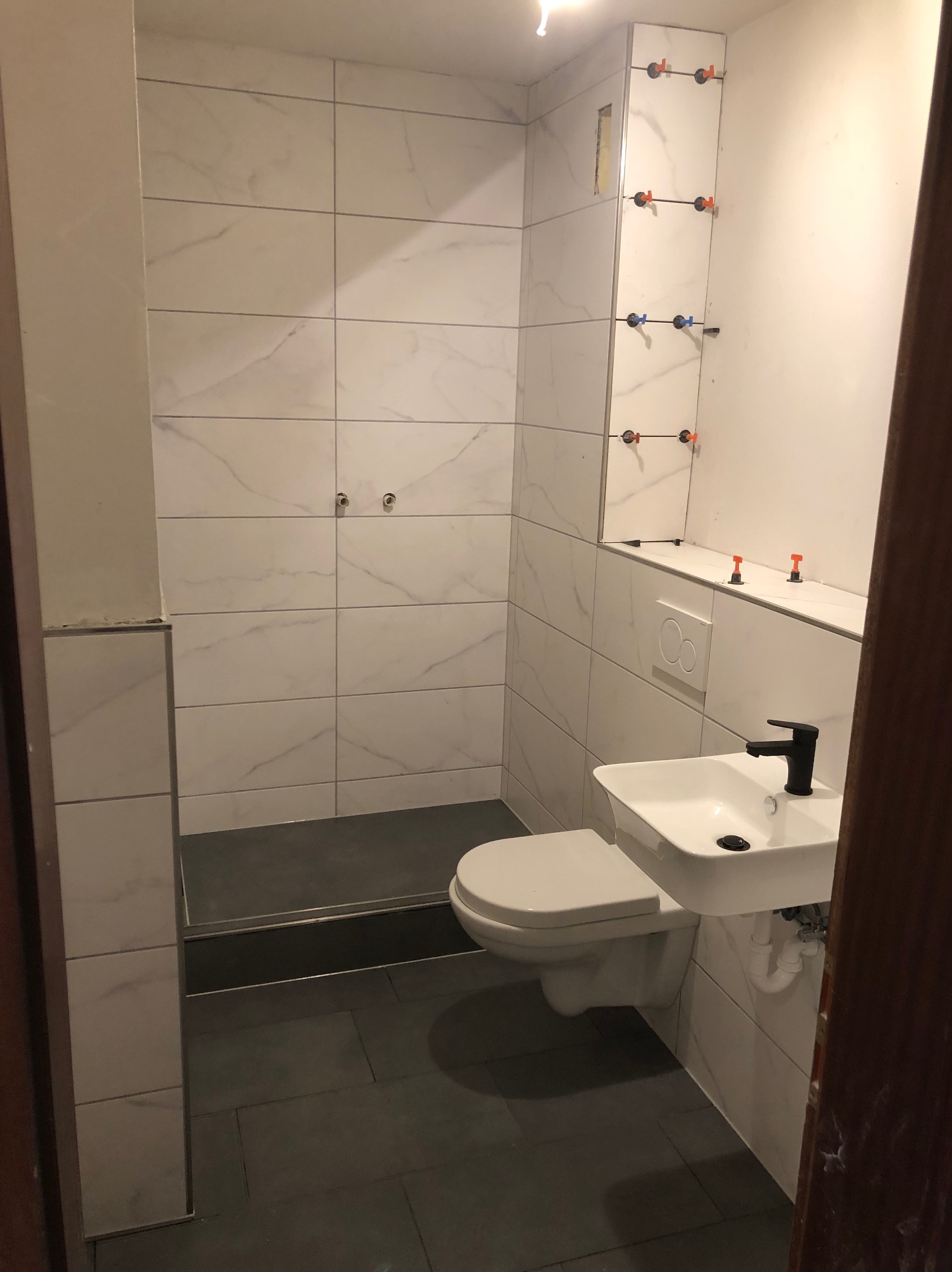 Nowoczesna łazienka w trakcie wykończenia: białe płytki imitujące marmur na ścianach, czarna armatura przy umywalce, widoczne elementy montażowe płytek.