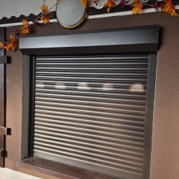 Ciemnobrązowa roleta zewnętrzna, częściowo opuszczona, zamontowana w oknie. Dekoracja jesienna nad roletą. Teksturowana ściana w odcieniach brązu.