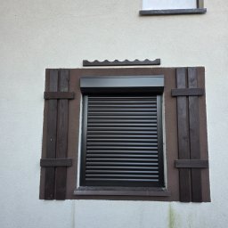 Zamknięta, czarna roleta zewnętrzna z drewnianymi okiennicami na elewacji budynku. Nad roletą dekoracyjny element imitujący dachówkę.