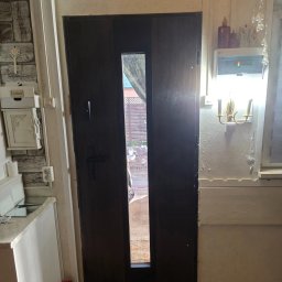 Ciemne drzwi zewnętrzne z wąskim przeszkleniem, widok na ogród. Po lewej skrzynka na listy, po prawej lampa ścienna. Drewniana podłoga, jasne ściany.