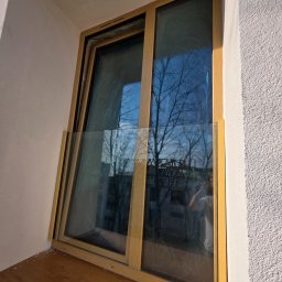 Okna i Drzwi Konrad Michalak - Nowe okno z drewnianą ramą, zabezpieczone folią, opiera się o ścianę w jasnym wnętrzu. Widoczne odbicie drzew na szybie.