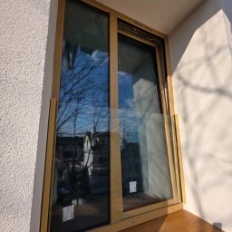 Okna i Drzwi Konrad Michalak - Nowe okno z drewnianą ramą, częściowo zabezpieczone folią, zamontowane w białej ścianie. W szybie odbija się błękitne niebo i korony drzew.