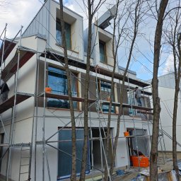 Okna i Drzwi Konrad Michalak - Budynek w trakcie prac wykończeniowych, widoczne rusztowania, okna zabezpieczone folią, pracownik na rusztowaniu. Ujęcie z dołu, drzewa na pierwszym planie.