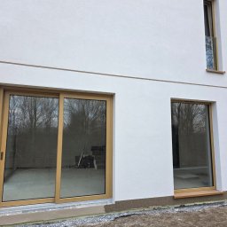 Okna i Drzwi Konrad Michalak - Nowoczesny dom z jasną elewacją i oknami z drewnianymi ramami. Duże okno przesuwne z widokiem na ogród. Minimalistyczny design.