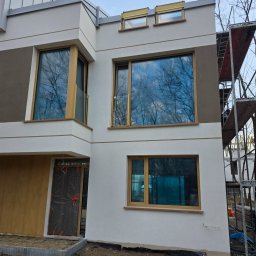 Okna i Drzwi Konrad Michalak - Nowoczesny dom z jasną elewacją i drewnianymi oknami, w tle rusztowanie. W oknach odbija się błękitne niebo i korony drzew.