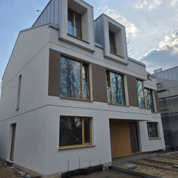 Okna i Drzwi Konrad Michalak - Nowoczesny dom jednorodzinny z oknami i drzwiami w Poddębicach. Elewacja w odcieniach bieli i szarości, okna z drewnianymi ramami. Widoczne rusztowania.