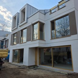 Okna i Drzwi Konrad Michalak - Nowoczesny budynek z dużymi oknami z drewnianymi ramami, odbijającymi drzewa i niebo. Widoczne detale wykończenia elewacji i otoczenie w trakcie prac.