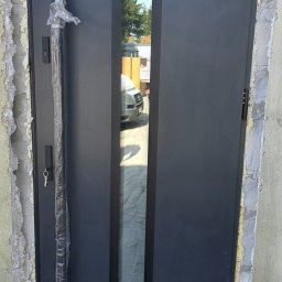 Okna i Drzwi Konrad Michalak - Grafitowe drzwi wejściowe z szybą, z długim uchwytem w folii ochronnej, osadzone w surowym otworze. Widoczne pianowanie i klucz w zamku.