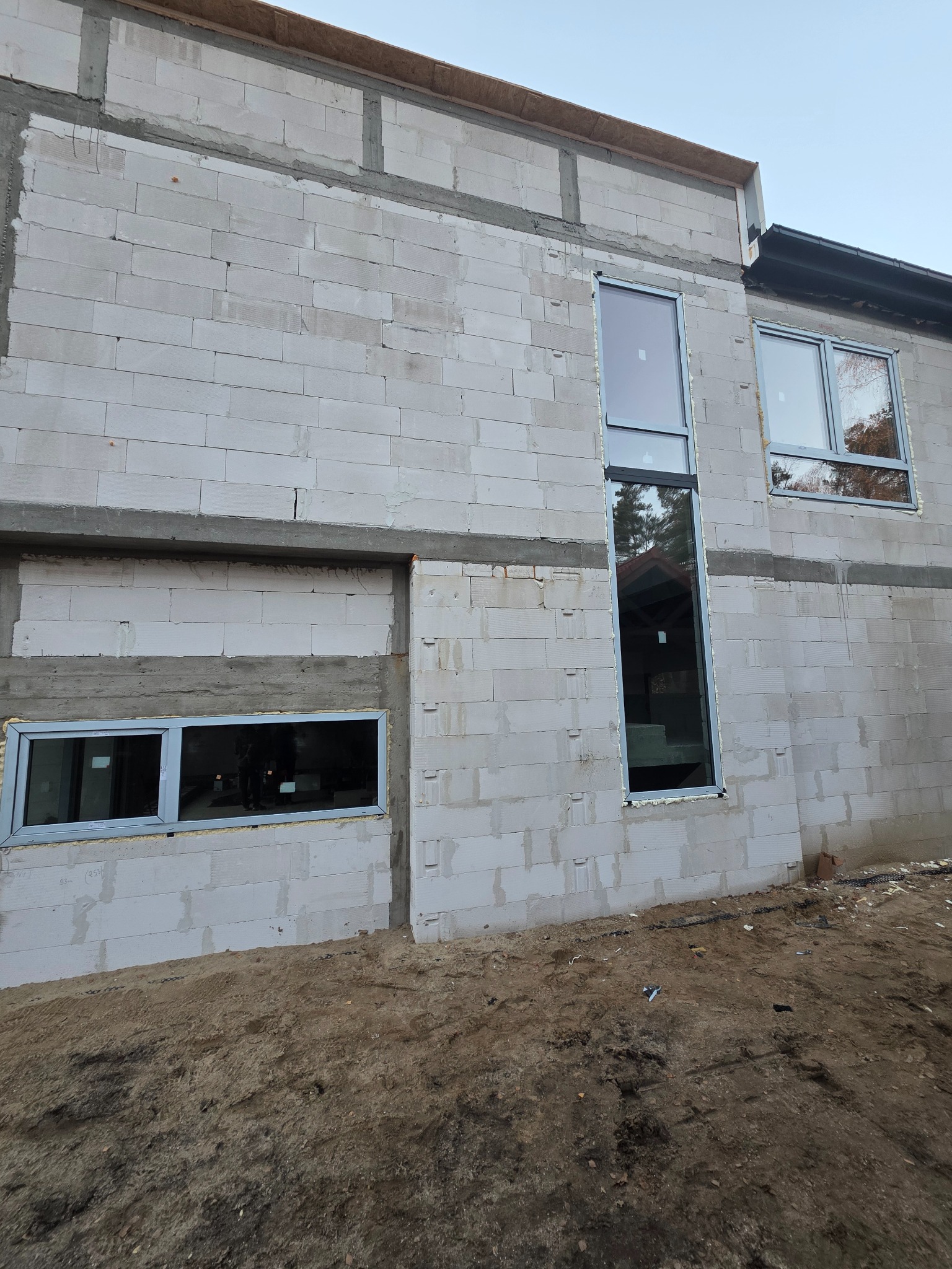 Surowy budynek z zamontowanymi oknami o różnych rozmiarach. Widoczne bloczki betonowe i piasek. Okna w szarych ramach, odzwierciedlają otoczenie.