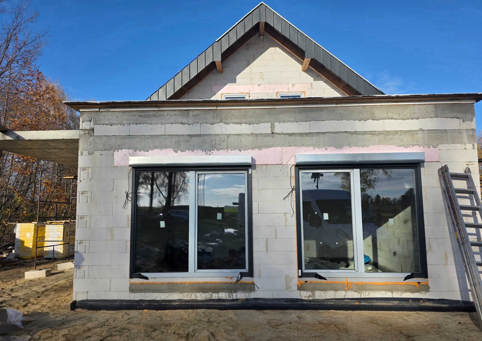 Montaż okien PCV w surowym budynku z szarej cegły. Widoczne okna z roletami zewnętrznymi, drabina i materiały budowlane. Słoneczny dzień, błękitne niebo.