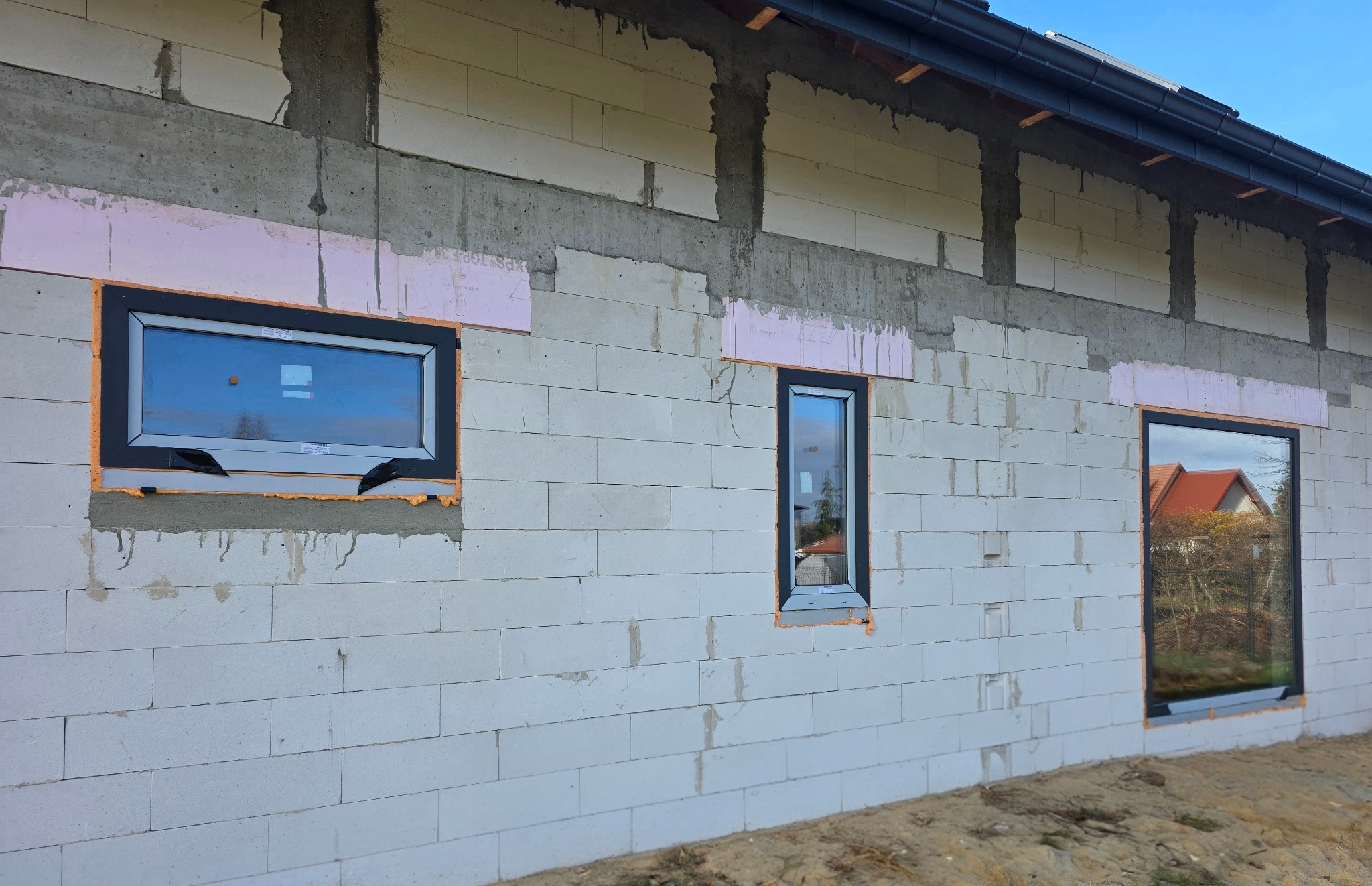 Montaż okien w surowym budynku z betonu komórkowego. Widoczne trzy okna z czarnymi ramami, zabezpieczone pianką montażową i styropianem. Etap prac wykończeniowych.