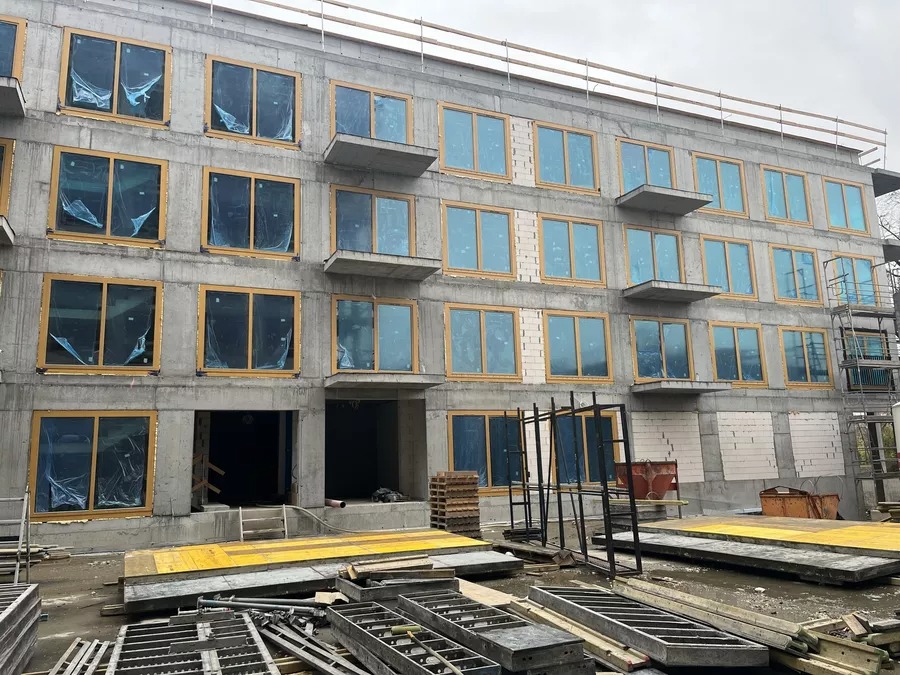 Montaż okien w nowym budynku w Poddębicach. Widoczne okna z żółtymi ramami, zabezpieczone folią, na tle surowego betonu. Plac budowy z materiałami i rusztowaniami.