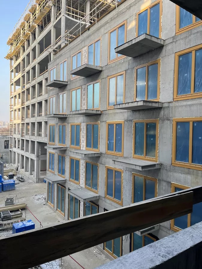 Wysoki budynek w trakcie budowy z zamontowanymi oknami z drewnianą ramą, zabezpieczonymi folią. Widok z góry na plac budowy i okolicę.