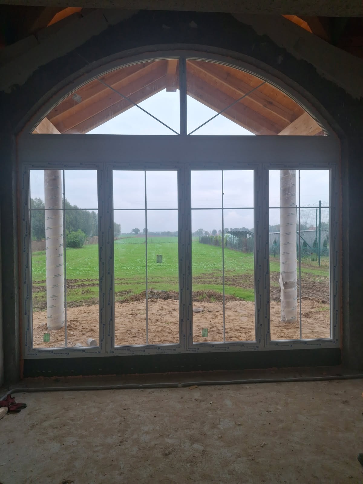 Nowe okno PCV z łukowym nadprożem w surowym wnętrzu. Widok na zielone pole przez szyby. Folia ochronna na słupku okna. Surowy beton i drewniana konstrukcja dachu.
