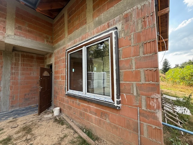 Białe okno PCV z brązową ramą, zamontowane w surowej ścianie z czerwonej cegły. Widoczny fragment brązowych drzwi wejściowych i teren budowy.