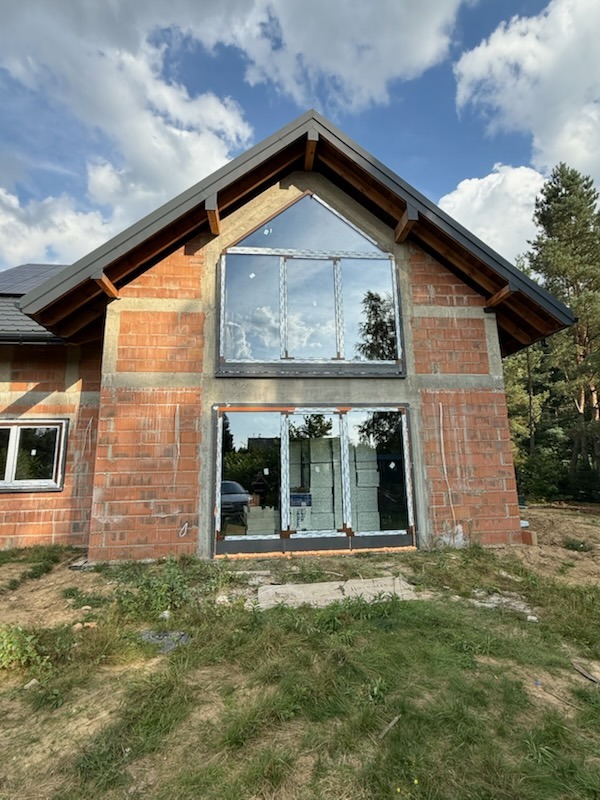 Surowy stan budynku z zamontowanymi oknami i drzwiami przesuwnymi. Widoczne cegły, okno trójkątne na górze i drzwi przesuwne na dole. W tle drzewa i niebo.