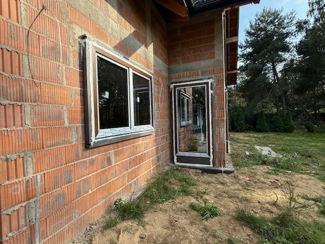 Montaż okna i drzwi zewnętrznych w surowym budynku z czerwonej cegły. Widoczne białe ramy okienne i brązowe ramy drzwiowe. Prace w toku, widoczne niedociągnięcia w murze.