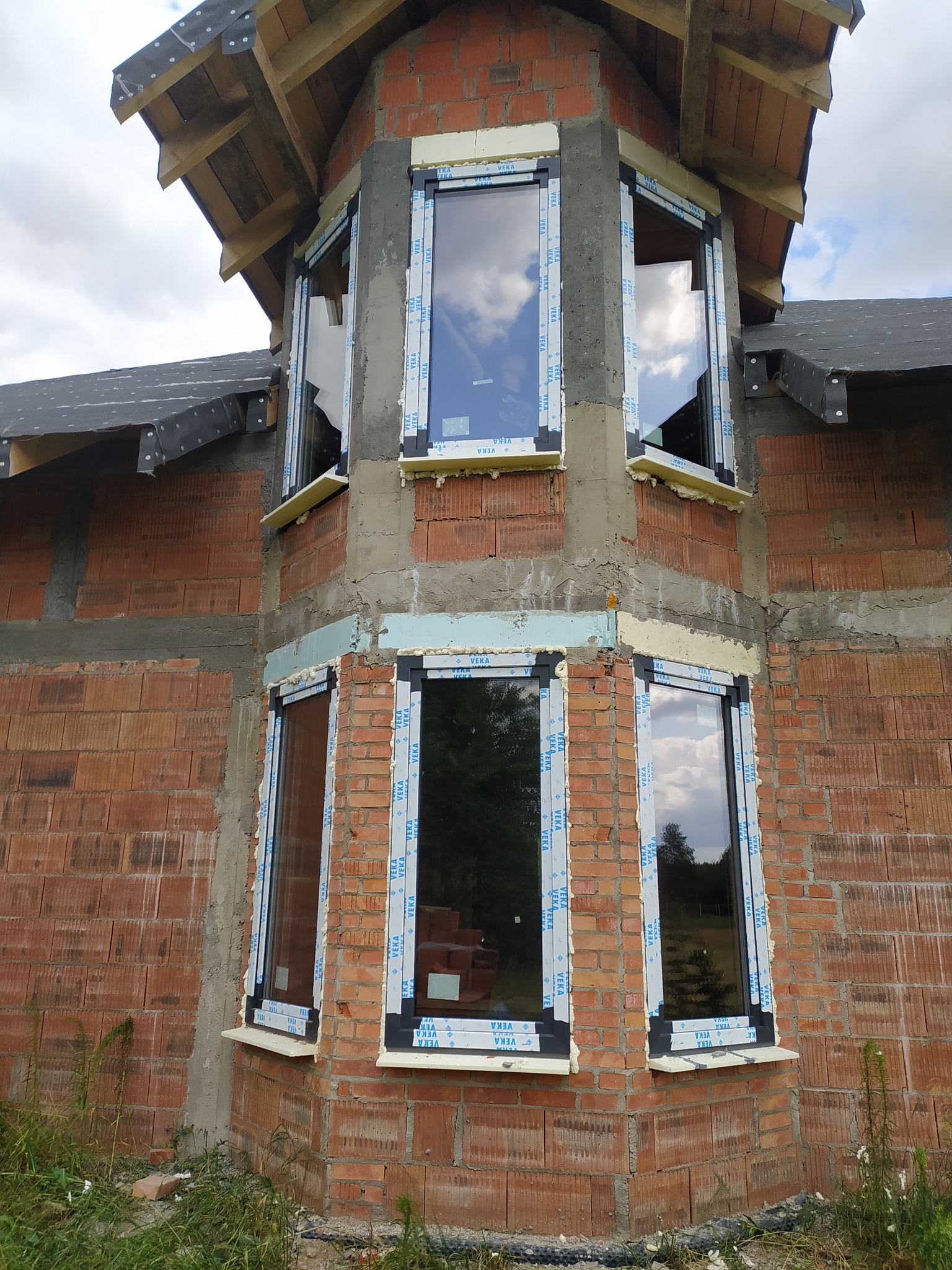 Montaż okien w surowym budynku z czerwonej cegły. Widoczne ramy okienne z pianką montażową. Okna odbijają niebo i drzewa. Budynek w trakcie budowy.