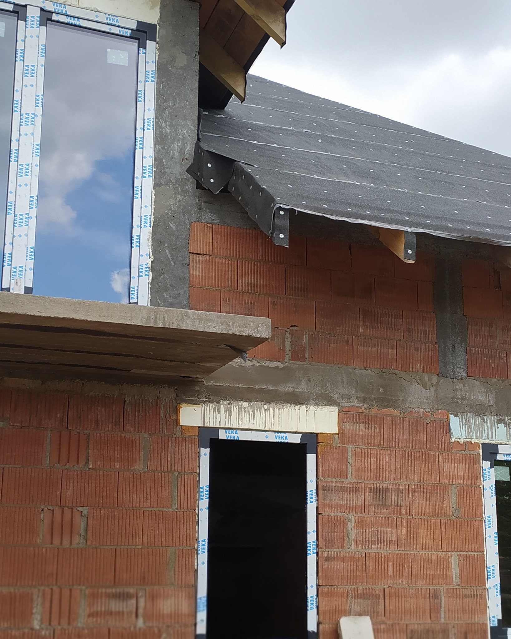 Surowy stan budynku z oknami i drzwiami w profilach VEKA, widoczne bloczki ceramiczne i fragment dachu z membraną. Etap montażu stolarki okiennej i drzwiowej.