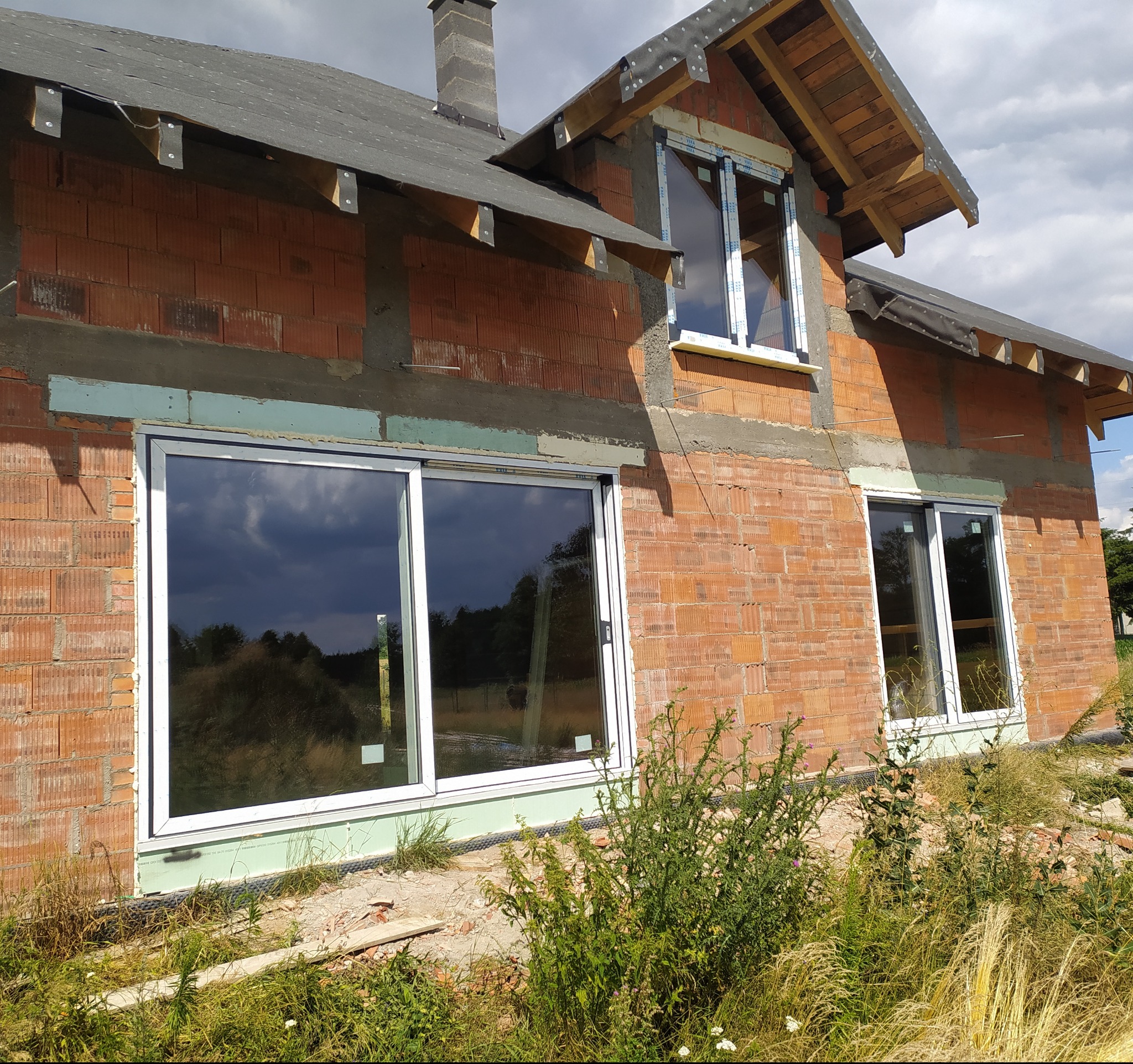 Dom w trakcie budowy z zamontowanymi oknami PCV. Widoczne okno przesuwne i okno na poddaszu. Budynek z czerwonej cegły.