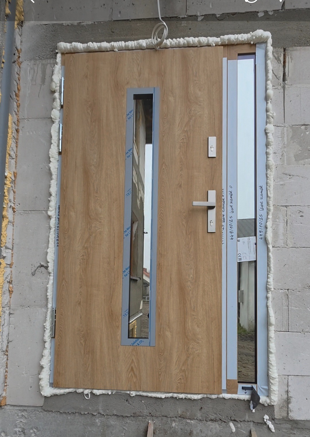 Montaż drzwi zewnętrznych z przeszkleniem i drewnopodobnym wykończeniem, uszczelnionych pianką montażową w surowym murze. Widoczny fragment elewacji budynku.