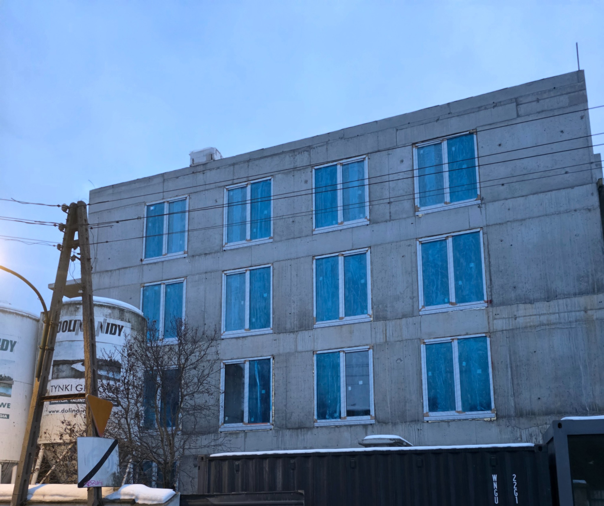 Surowy budynek wielorodzinny w trakcie budowy, z zamontowanymi oknami zabezpieczonymi folią, widoczne silosy z tynkiem gipsowym i kontener budowlany.