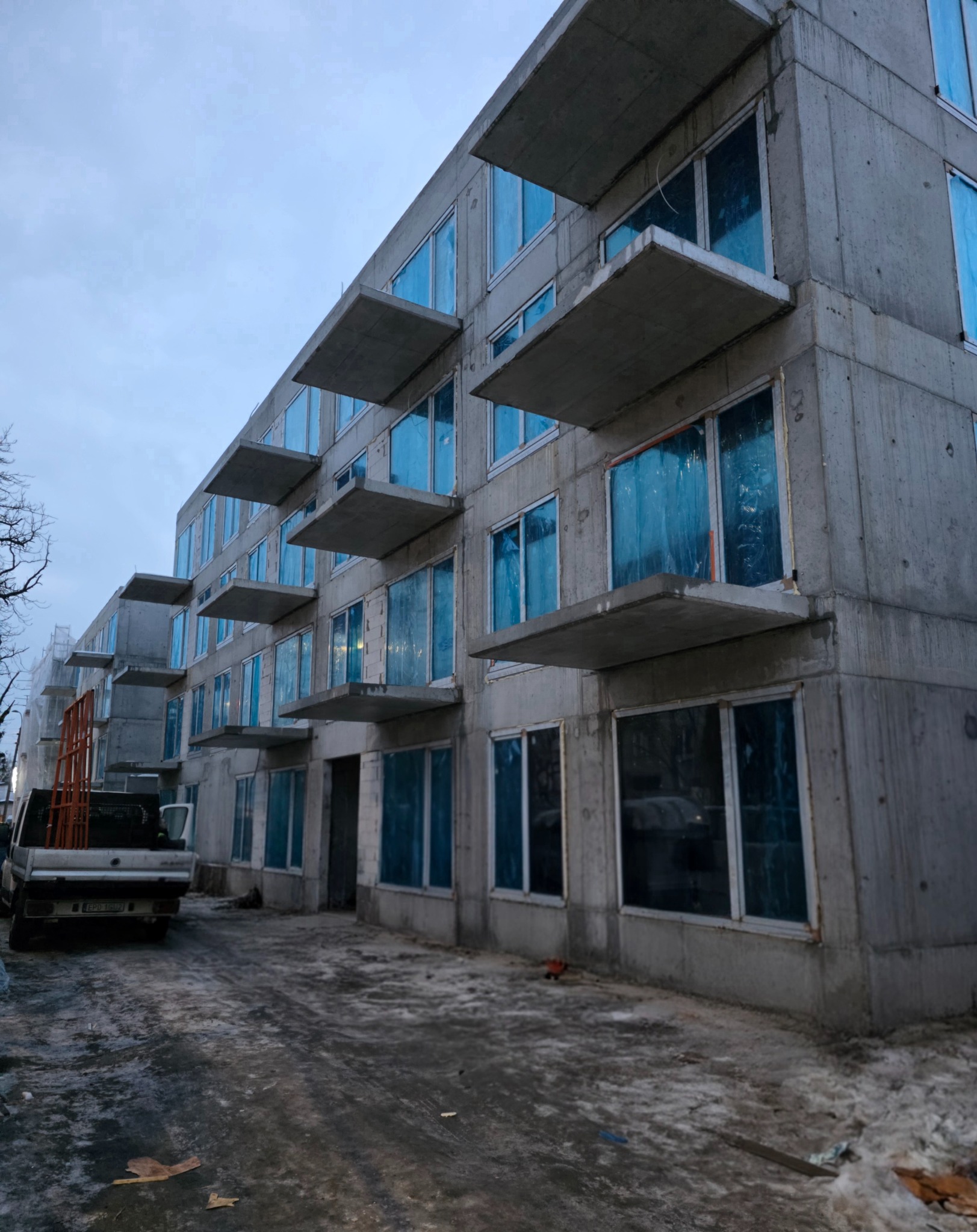 Surowy budynek wielorodzinny z betonowymi balkonami i oknami zabezpieczonymi folią, w tle zaparkowany samochód dostawczy z elementami montażowymi.