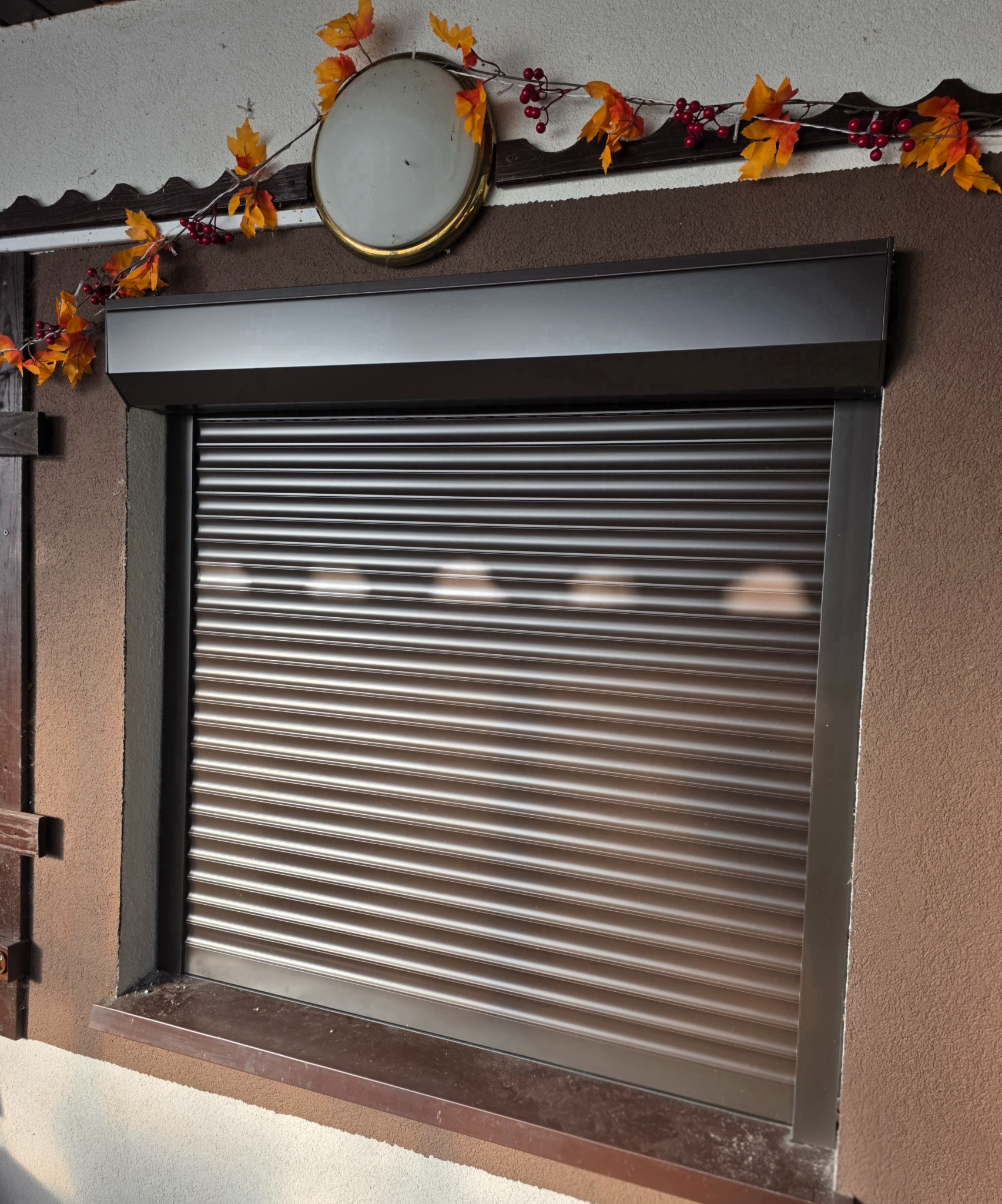 Ciemnobrązowa roleta zewnętrzna, częściowo opuszczona, zamontowana w oknie. Dekoracja jesienna nad roletą. Teksturowana ściana w odcieniach brązu.